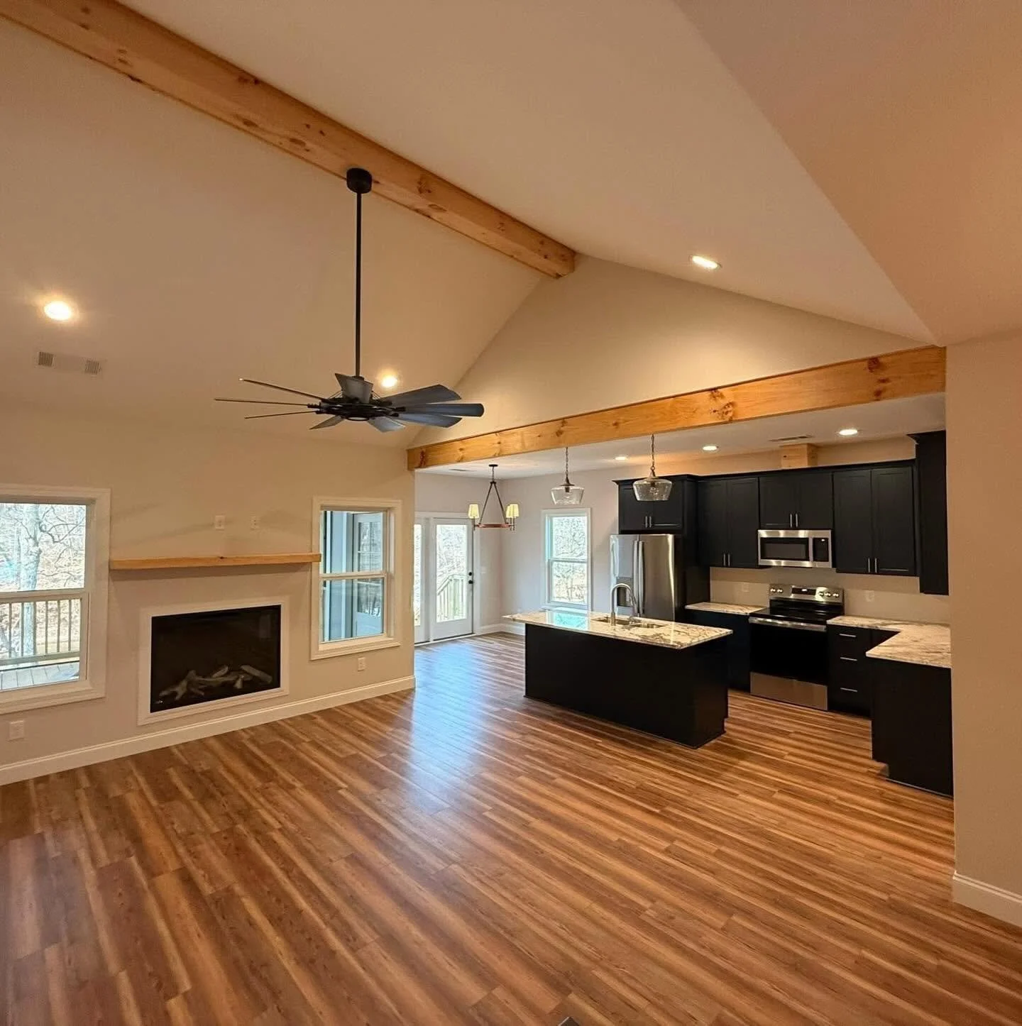 Open living room and kitchen with hardwood floors, black cabinets, stainless steel appliances, a fireplace beneath two windows, a ceiling fan, and a tray ceiling with wood beams.