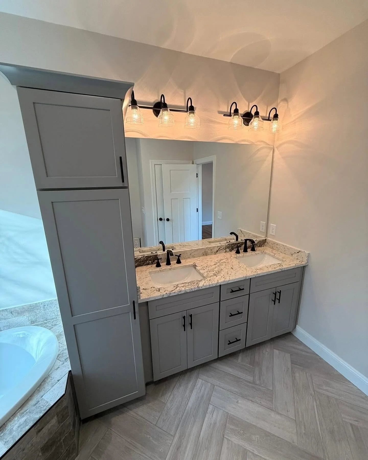 Bathroom vanity with a granite countertop, double sinks, and black fixtures. Large mirror with six light fixtures above it. Light-colored walls and flooring, with a doorway leading to another room.