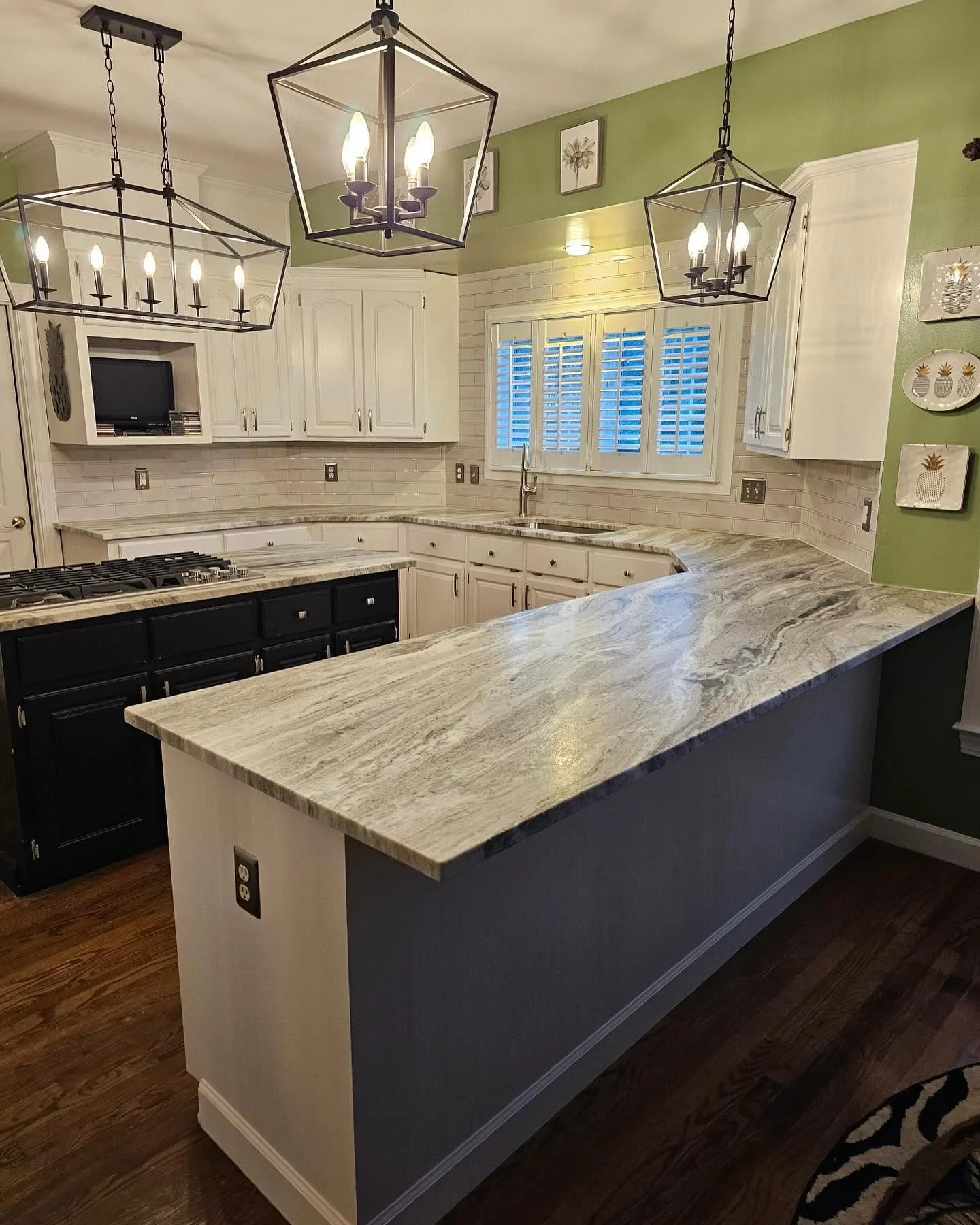 Kitchen with white cabinets, a marble countertop, black lower cabinets, and a green wall. Hanging geometric black chandeliers, a window with shutters, and wall art with pineapples.