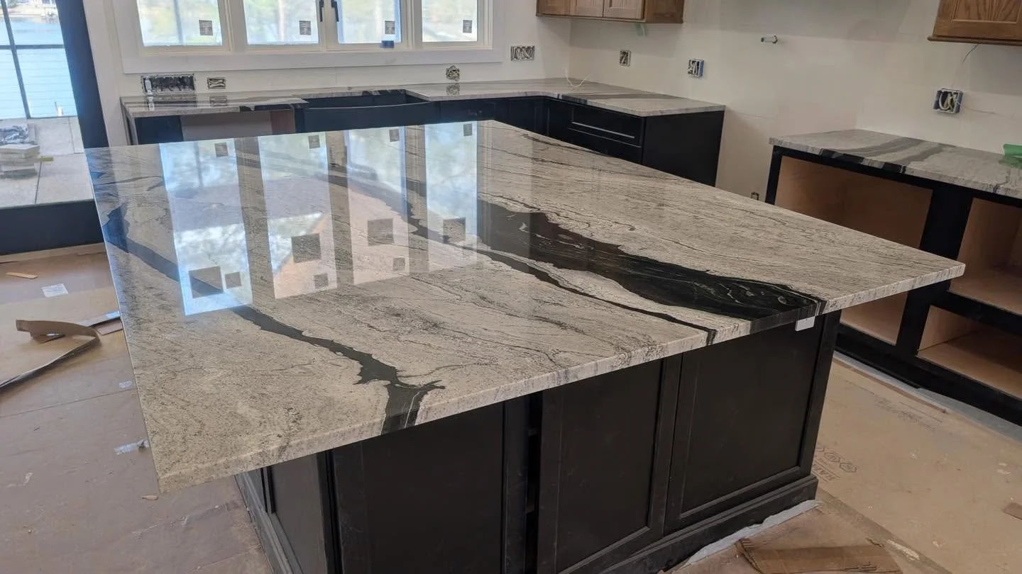 A kitchen island with a polished granite countertop with black and beige veining in a new or under-construction kitchen. There are no appliances or fixtures attached yet, and surrounding cabinetry is missing doors.