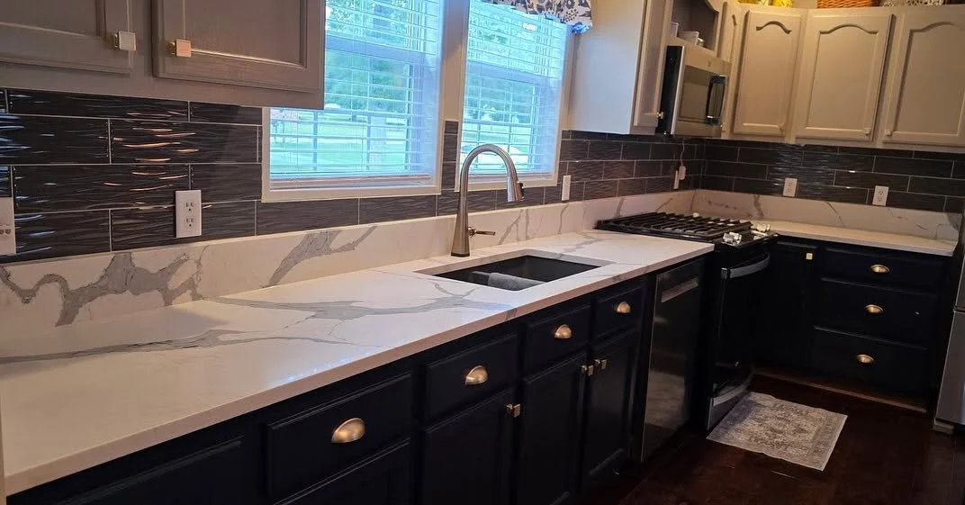 Modern kitchen with black lower cabinets, white upper cabinets, marble countertop with a sink and a stove, black backsplash tiles, and two windows with blinds.
