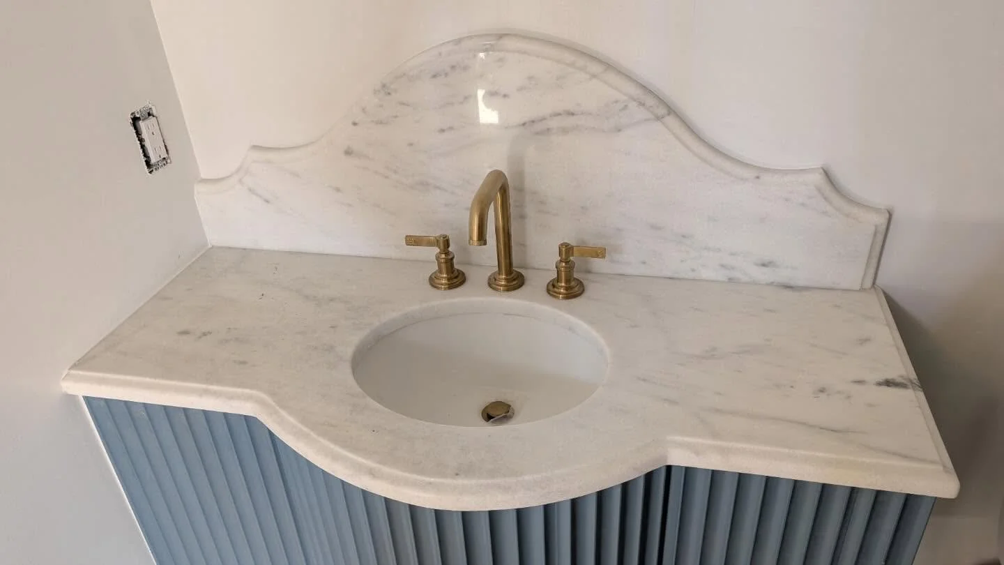 Marble bathroom vanity with a curved edge, gold faucet, two gold handles, a marble backsplash, and a blue cabinet with vertical ridges.