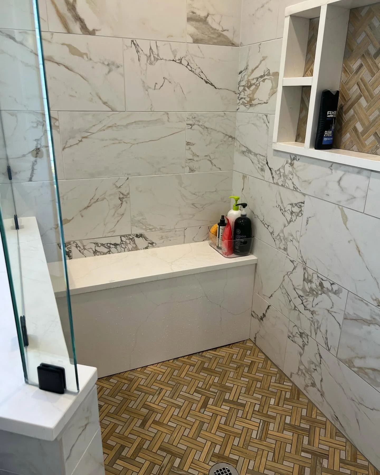 A shower area with white marble wall tiles featuring gray veining and a built-in shelf holding toiletry bottles. The floor has a geometric wood-patterned tile design, and a part of a glass shower door is visible on the left side.