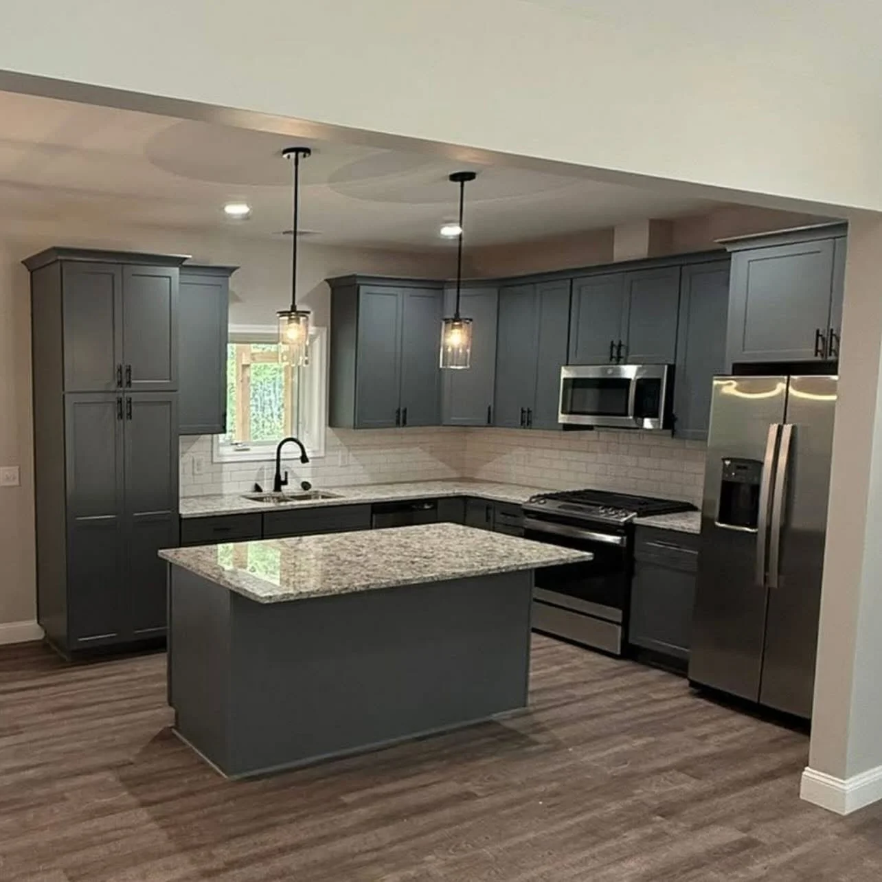Modern kitchen with grey cabinets, granite countertops, white subway tile backsplash, and stainless steel appliances, including a refrigerator, microwave, and oven, illuminated by two pendant lights.