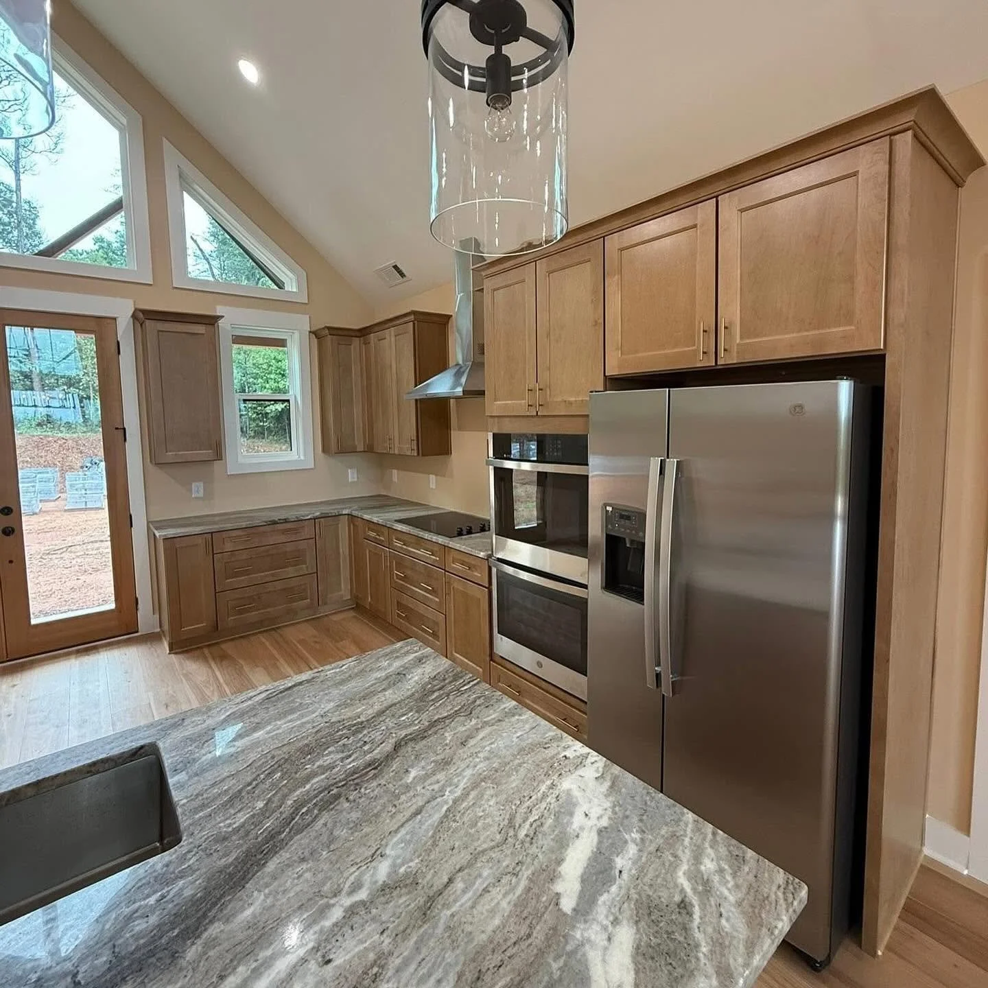 Modern kitchen with wooden cabinets, stainless steel refrigerator and oven, granite countertop, and large windows with a view of trees outside.