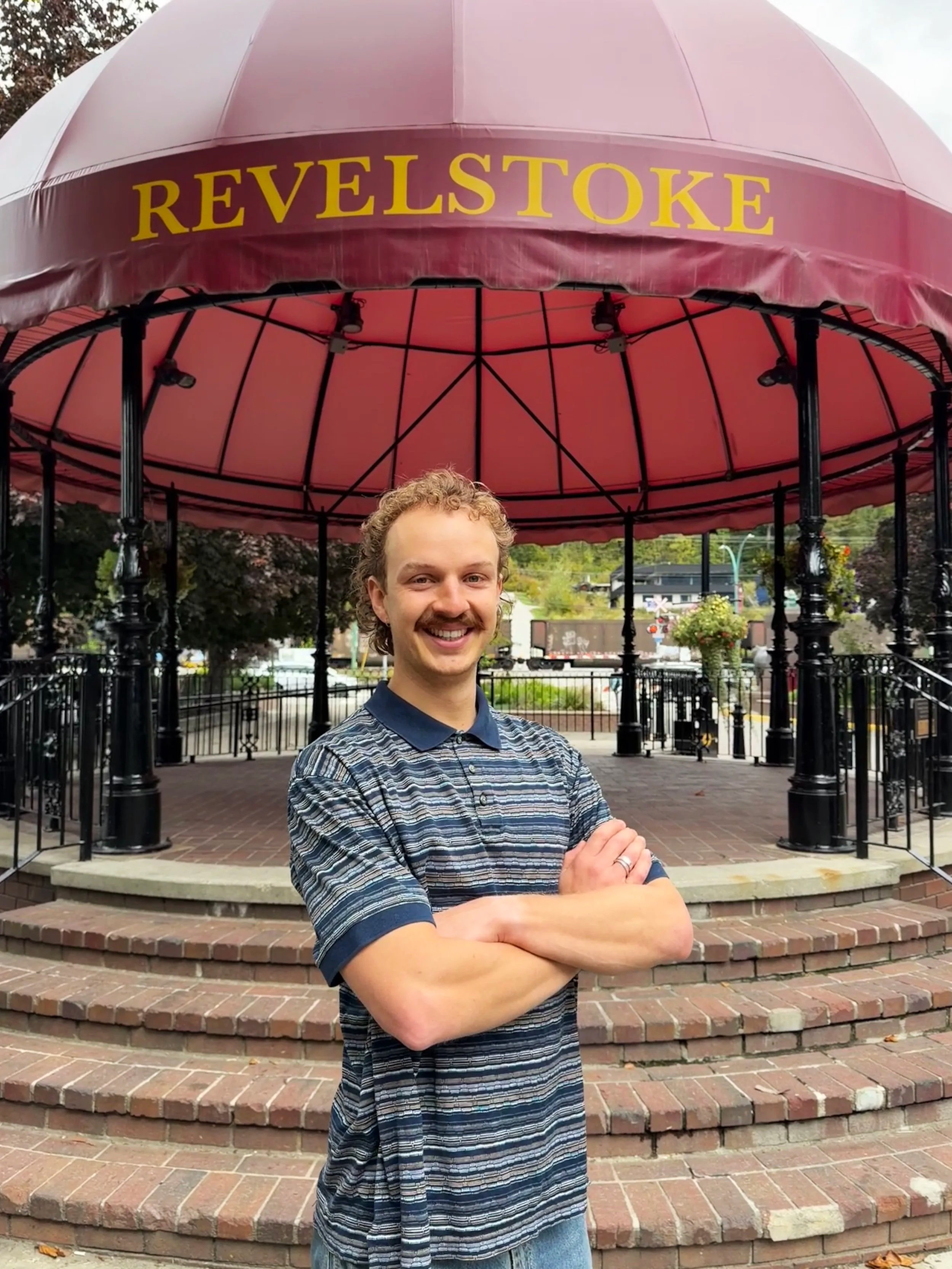 A man with curly hair and a mustache smiling with arms crossed, standing in front of a bandstand with a pink canopy labeled 'Revelstoke' in yellow, in a park setting during daytime.