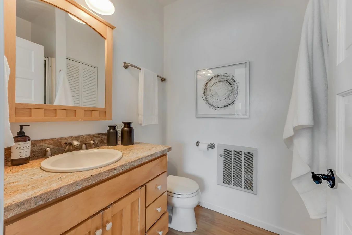 Bathroom with wooden vanity, granite countertop, oval sink, soap dispenser, toothbrush holder, mirror, white walls, framed artwork, towel on rack, hand towel on door, vent on wall, toilet.
