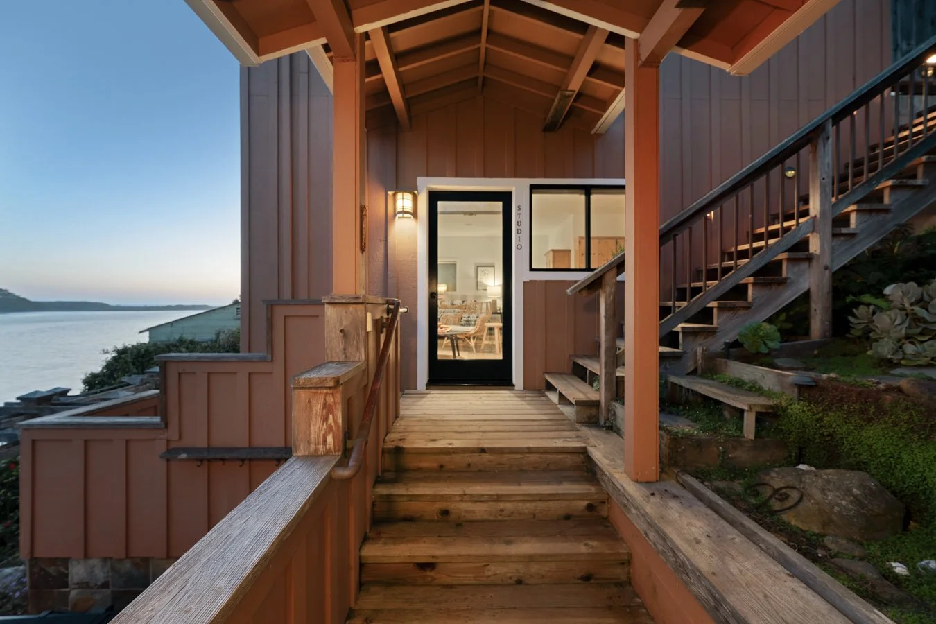 Wooden porch leading to a front door with a view of a body of water and an outdoor landscape at sunset.