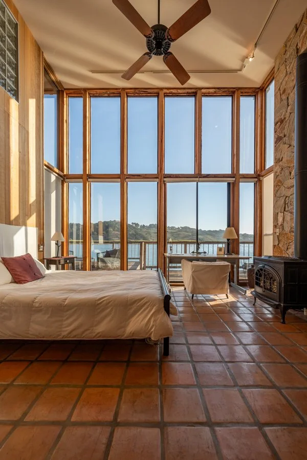 A bedroom with terracotta tile floor, large floor-to-ceiling windows overlooking a lake, a bed with white bedding, two nightstands with lamps, a ceiling fan, a wood stove, and an outdoor deck with seating.
