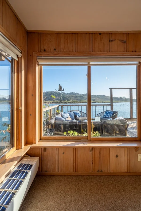 View of a wooden deck with outdoor furniture overlooking a lake, seen through large window in a room with wood-paneled walls.