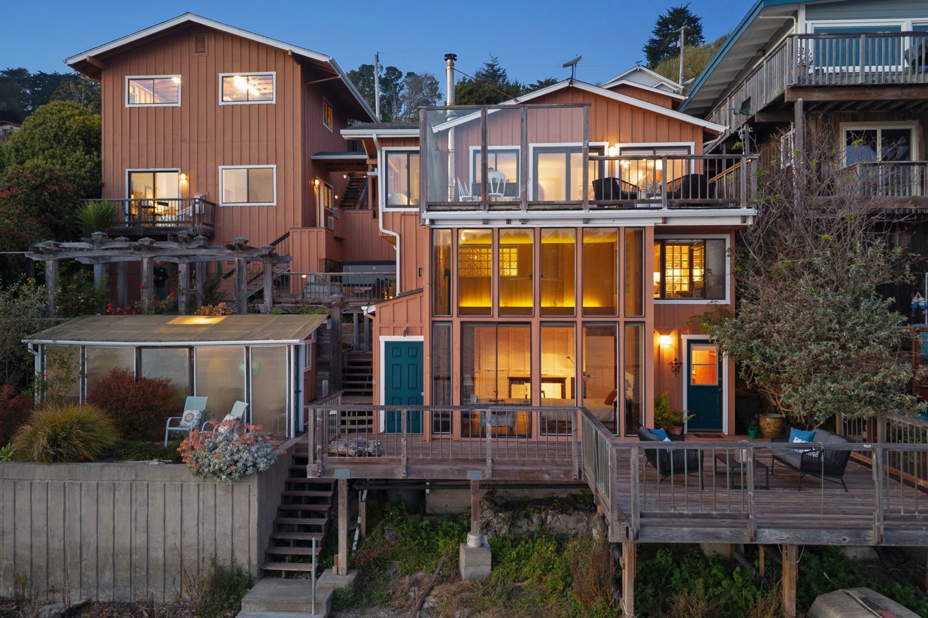 Multi-story house with large glass windows and multiple decks illuminated by interior lights during evening, surrounded by trees and neighboring houses