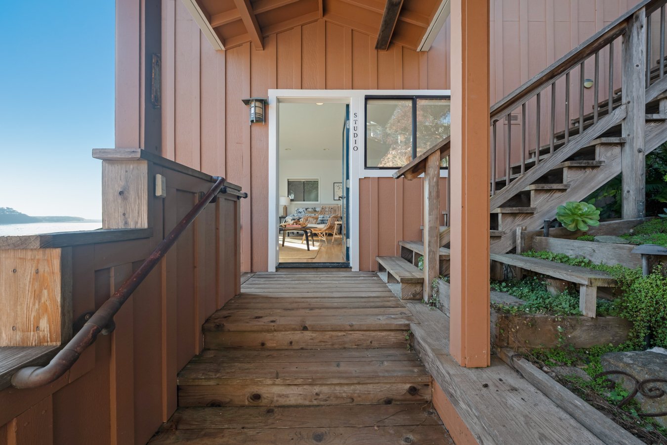 View of the patio entrance to a building with wood siding, stairs on the right, and a glass door leading into a room labeled 'STUDIO'. The room has a couch and interior decor visible through the door.