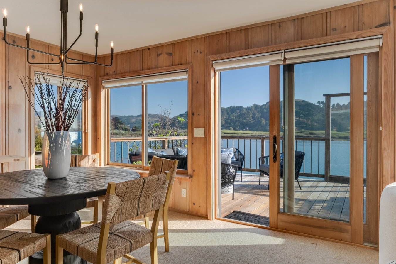 A dining room with wooden walls and large windows overlooking a water body and green hills, with a sliding glass door leading to a deck with outdoor seating and a railing.