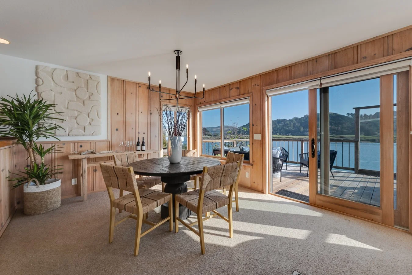 Dining room with a round wooden table, six chairs, a chandelier, and large sliding glass doors leading to a balcony overlooking a lake and hills.