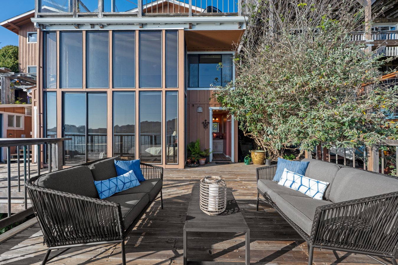 Outdoor wooden deck with two black outdoor sofas with blue and white pillows, a black coffee table, a large potted tree, and an exterior of a house with large glass windows and a secondary entrance.