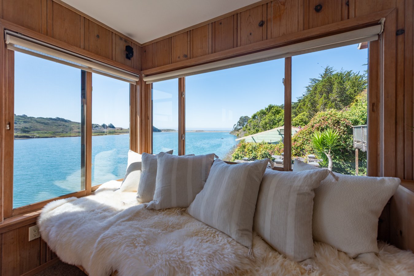Cozy window seat with white pillows and a fluffy throw blanket overlooking water and greenery outside.