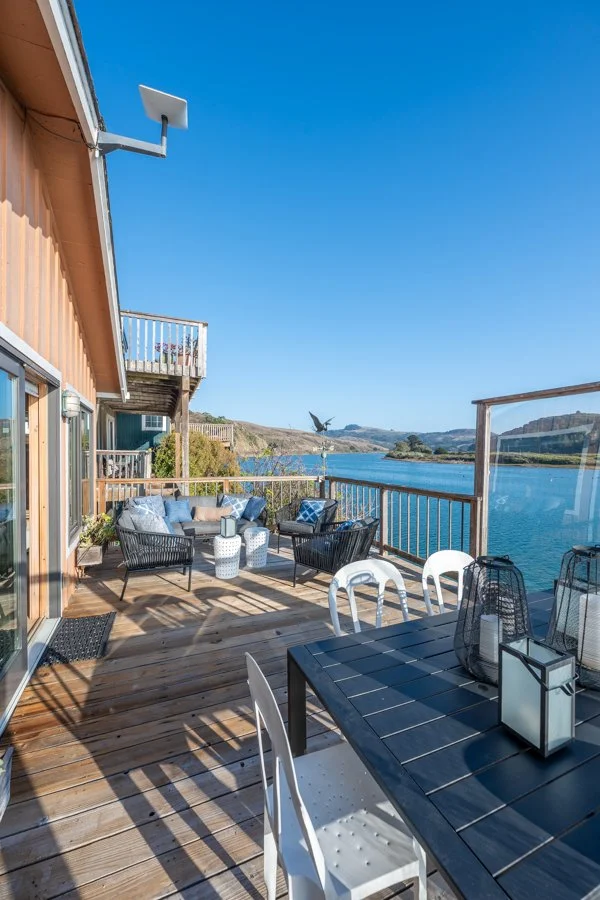 A wooden deck overlooking water with outdoor seating, a dining table with chairs, and a railing, under a clear blue sky.