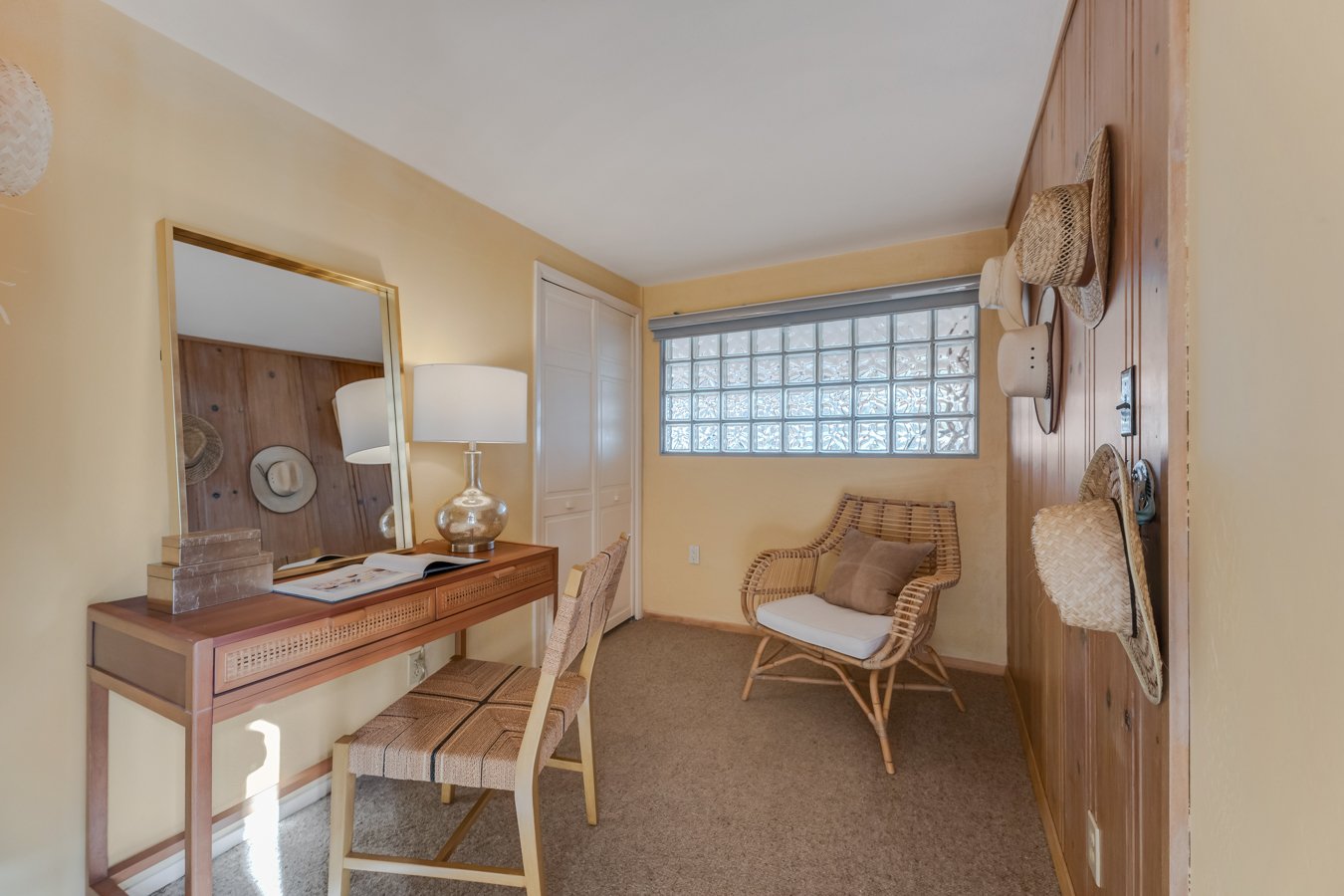 A cozy room with a wooden desk, mirror, lamp, and chair, decorated with straw hats on the wall and a wicker chair with pillows, illuminated by natural light from a glass-block window.