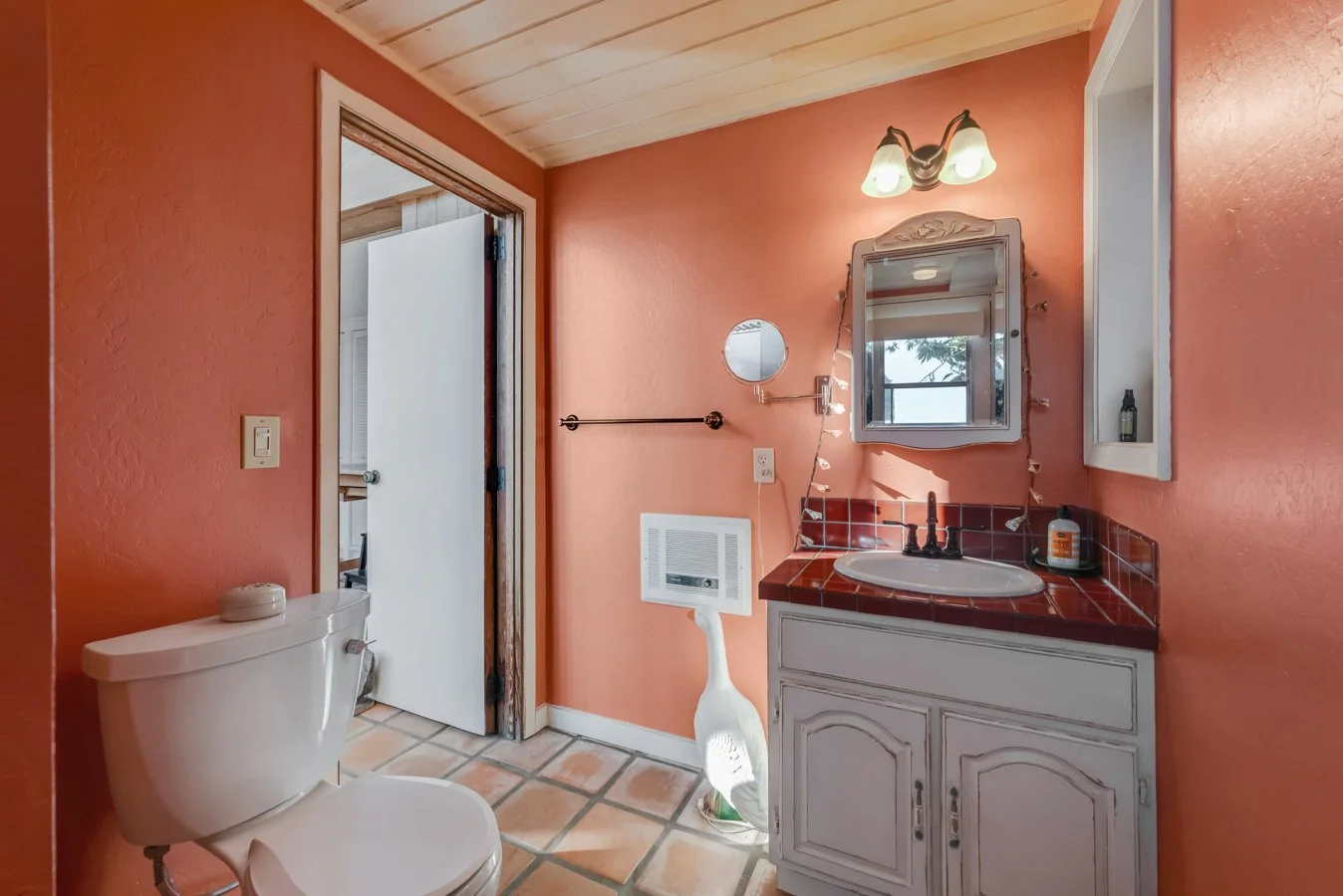 Bathroom with peach walls, white toilet, red tiled sink counter, mirror with decorative frame, small window, mounted light fixture, and a door leading to another room.