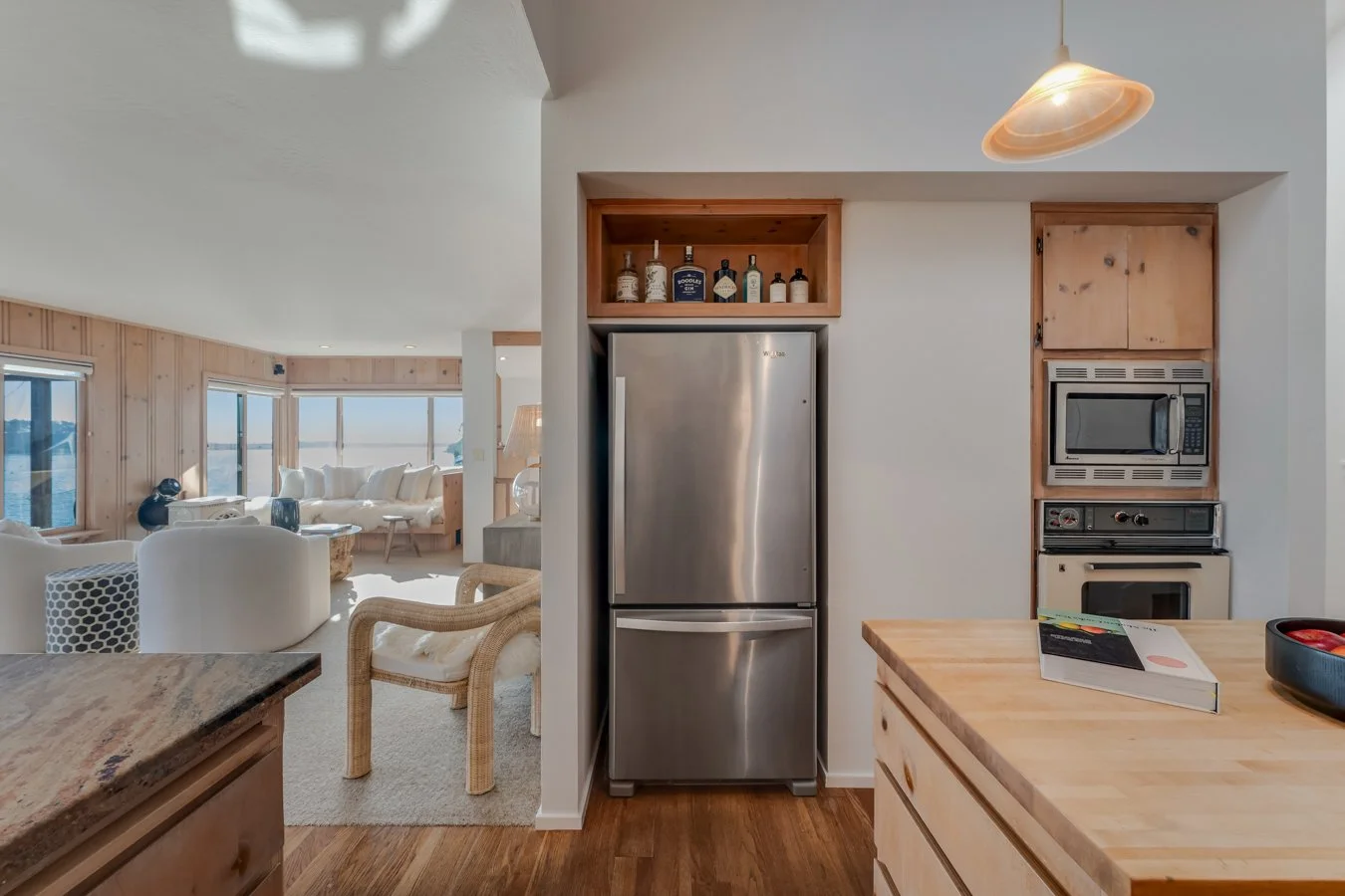 Kitchen with stainless steel refrigerator, microwave, oven, wooden cabinets, and a wooden kitchen island. Visible in the background is a living room with white furniture and large windows showing an ocean view.