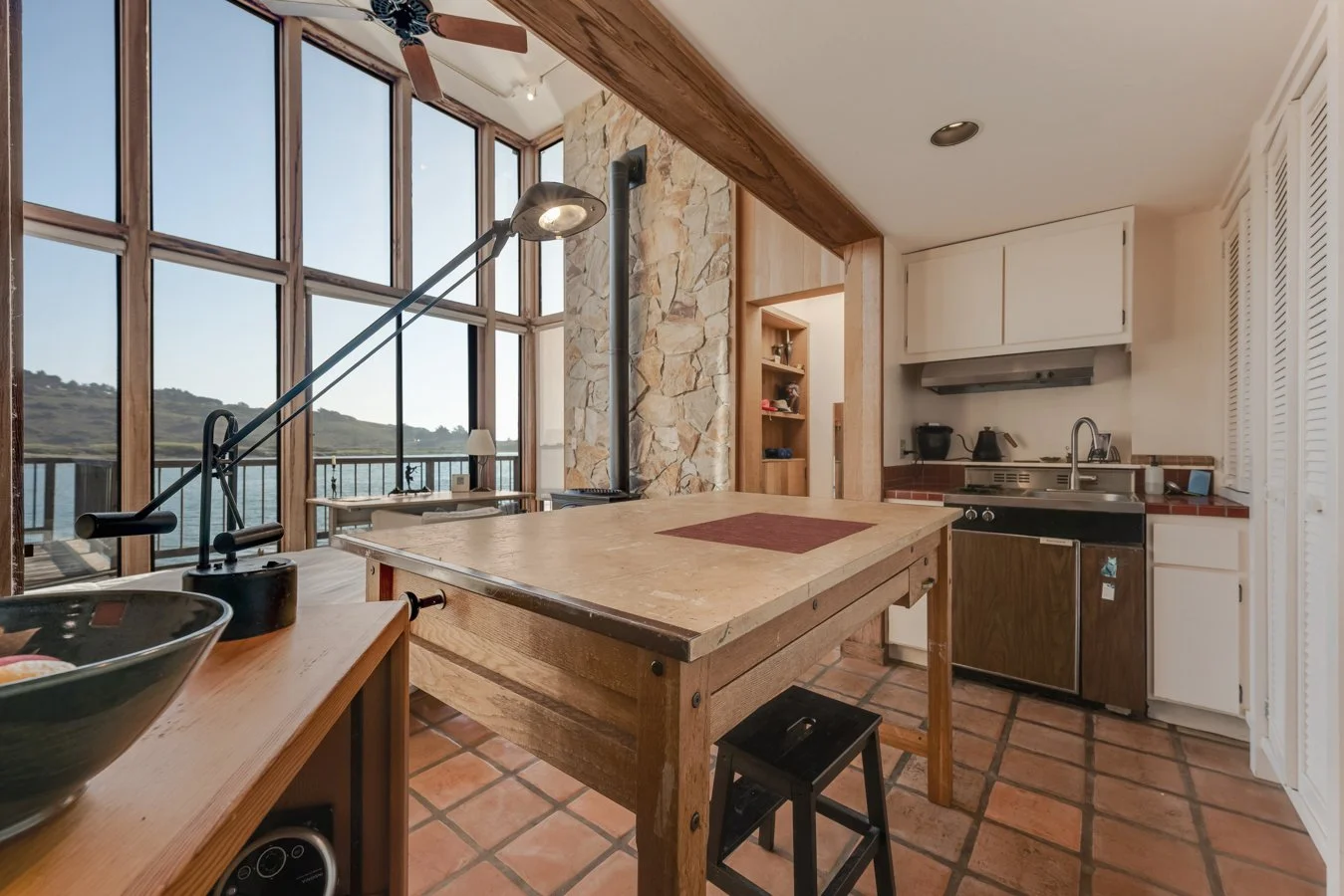 Kitchen with large windows overlooking a lake, wooden table, stove, sink, and cabinets.