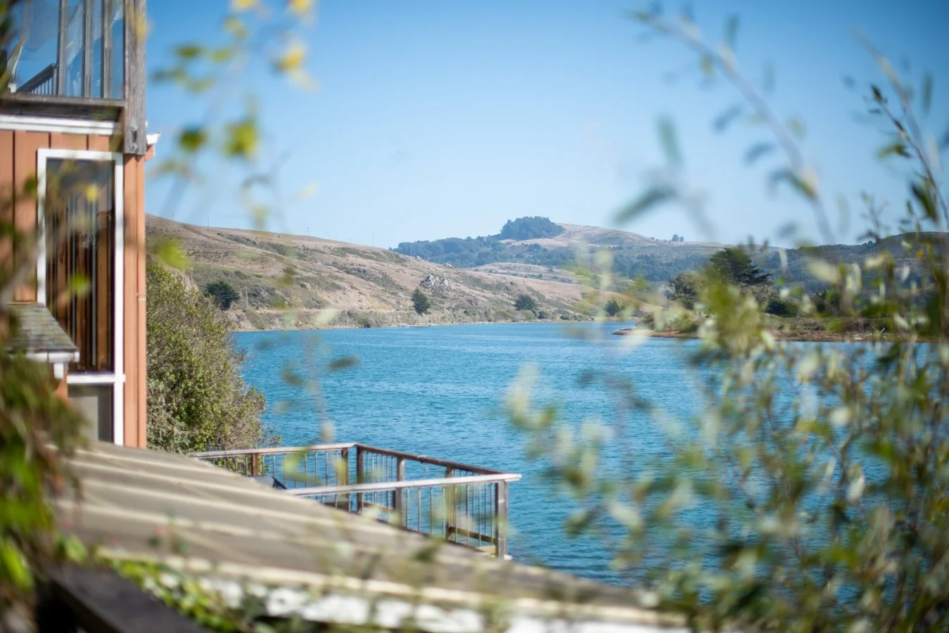 View of a lake surrounded by hills and trees, seen from a wooden balcony with railing, with a partially visible building on the left.