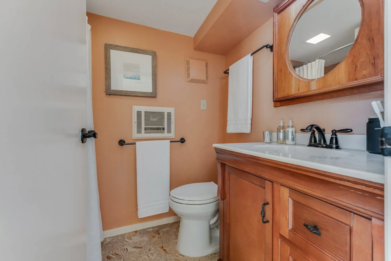 A small bathroom with peach-colored walls, a white toilet, a wooden vanity with a white countertop, a mirror, and various toiletries. There is a towel hanging on a bar and a framed picture above the toilet.