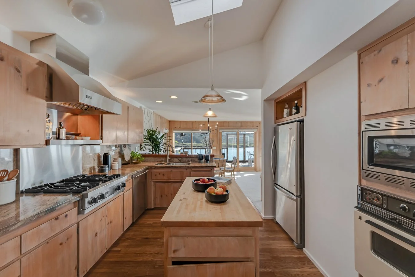 Modern kitchen with wooden cabinets, a central island with apples, stainless steel appliances, and a dining area with large windows and a lake view.