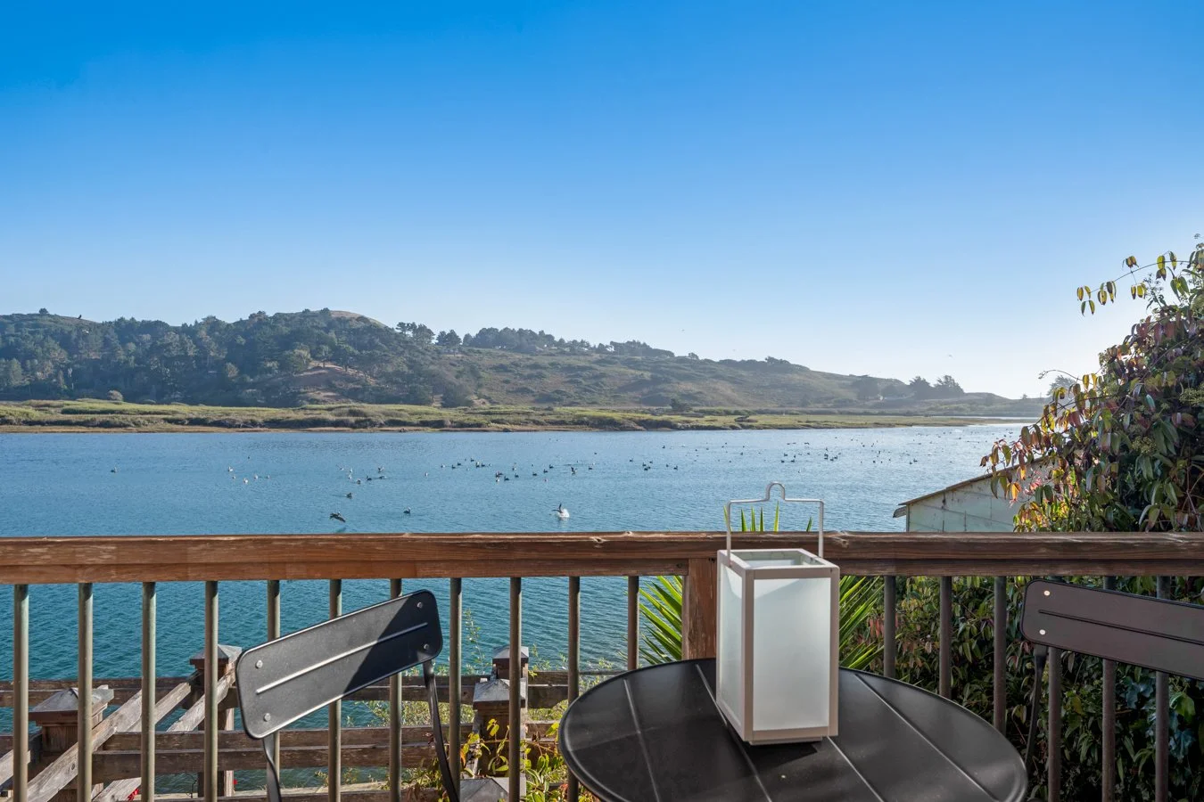 A balcony with a black table and chairs overlooking a serene lake with birds, green hills, and a clear blue sky.