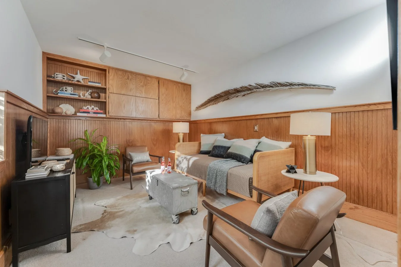 Living room with wooden paneling, beige carpeting, mid-century modern armchairs, a sofa with pillows, a flat-screen TV, wooden shelves with decorative items, side lamps, a green potted plant, a cowhide rug, and wall art resembling a large feather.