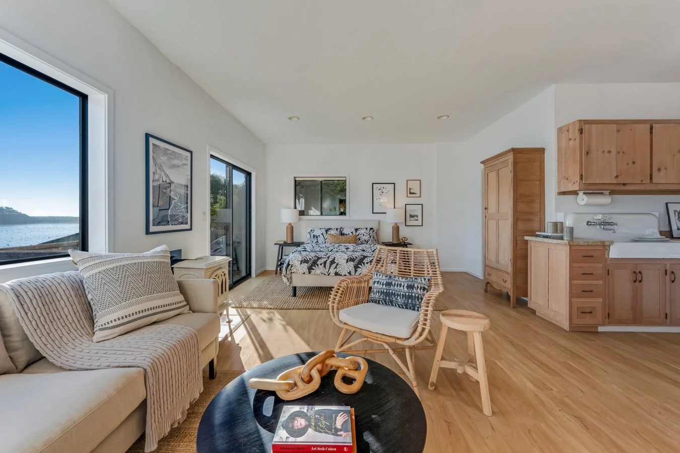 Open-concept living room and kitchen with large windows, a beige sofa, a black coffee table with decorative chain links, wicker armchair, bed with leaf-patterned bedding, and wooden kitchen cabinets.