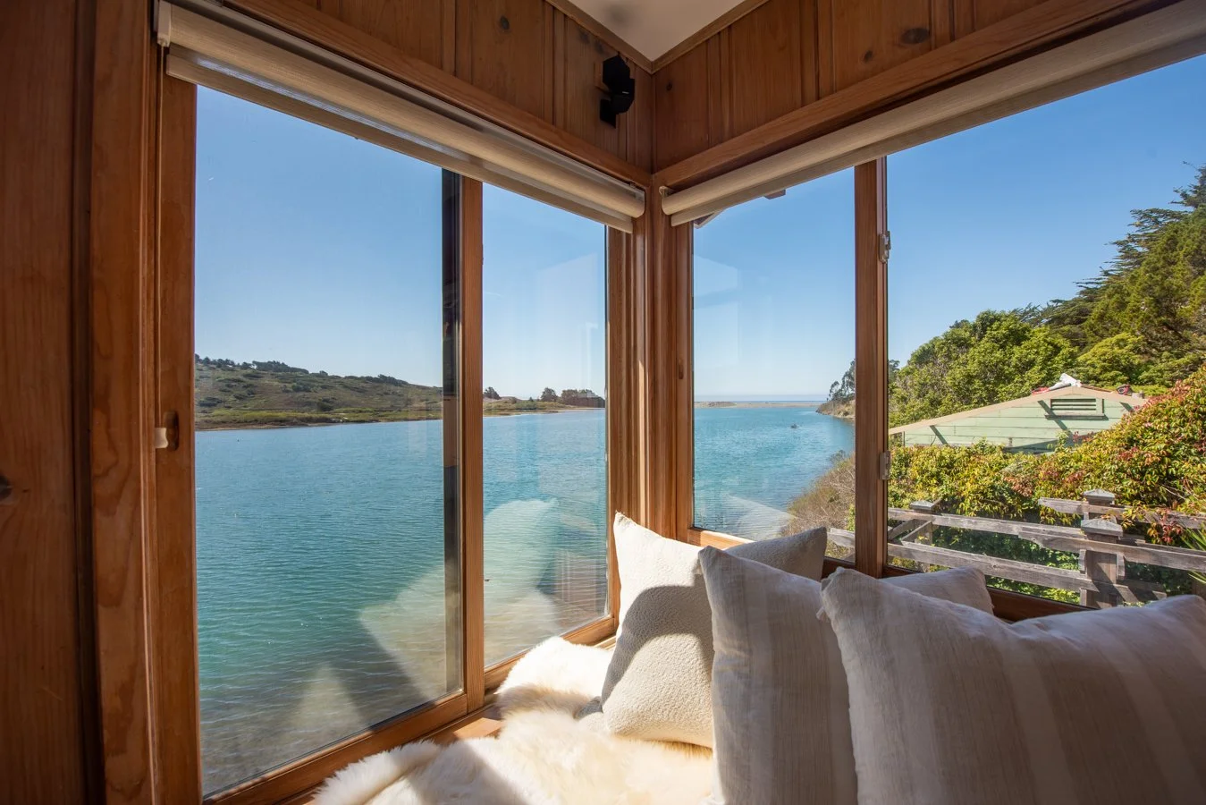 Cozy corner with large windows overlooking a body of water, with pillows and a furry blanket inside a wooden room.