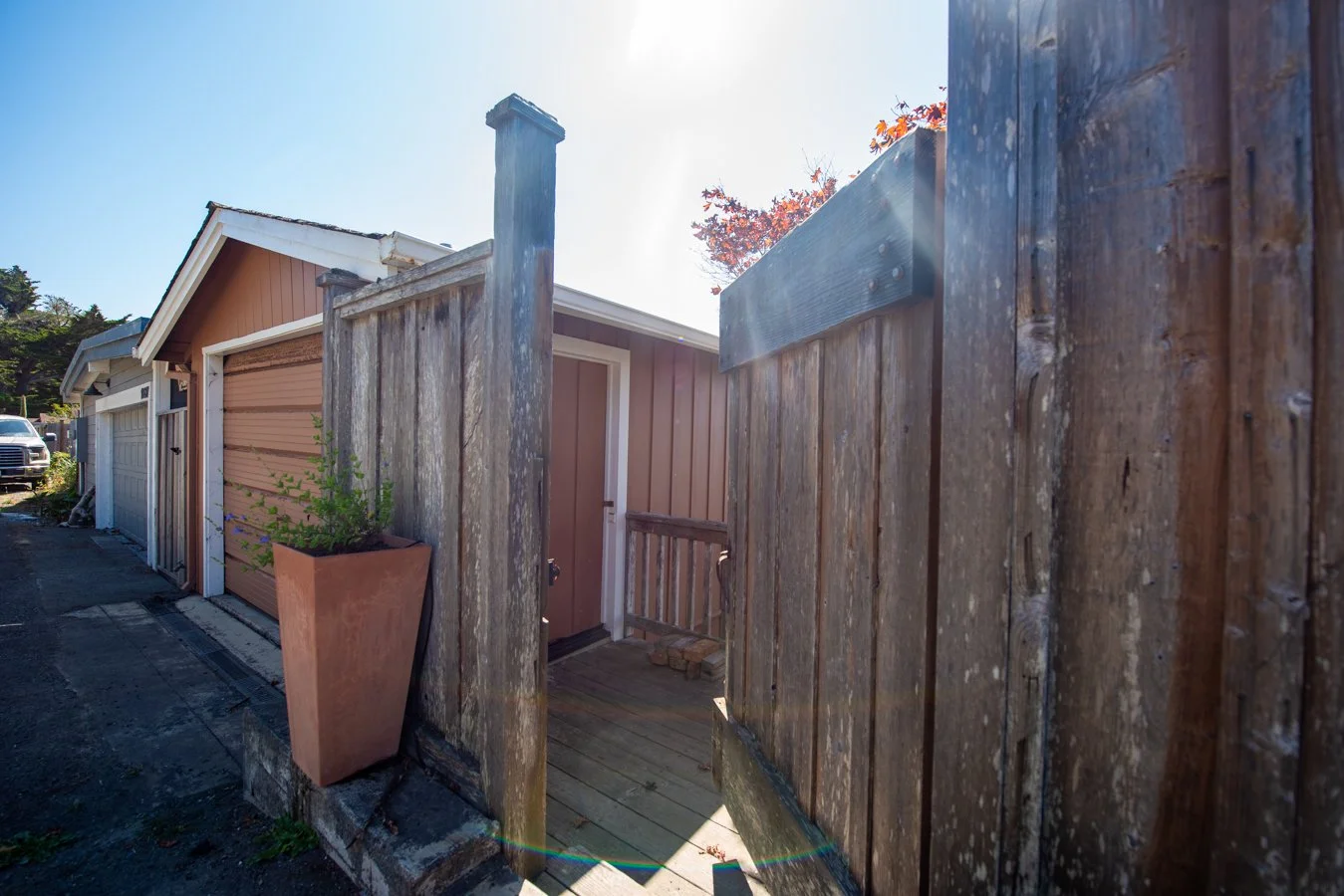 View of a fenced area with a small brown shed and a sidewalk with a potted plant, parked cars, and garages in the background, taken on a sunny day.