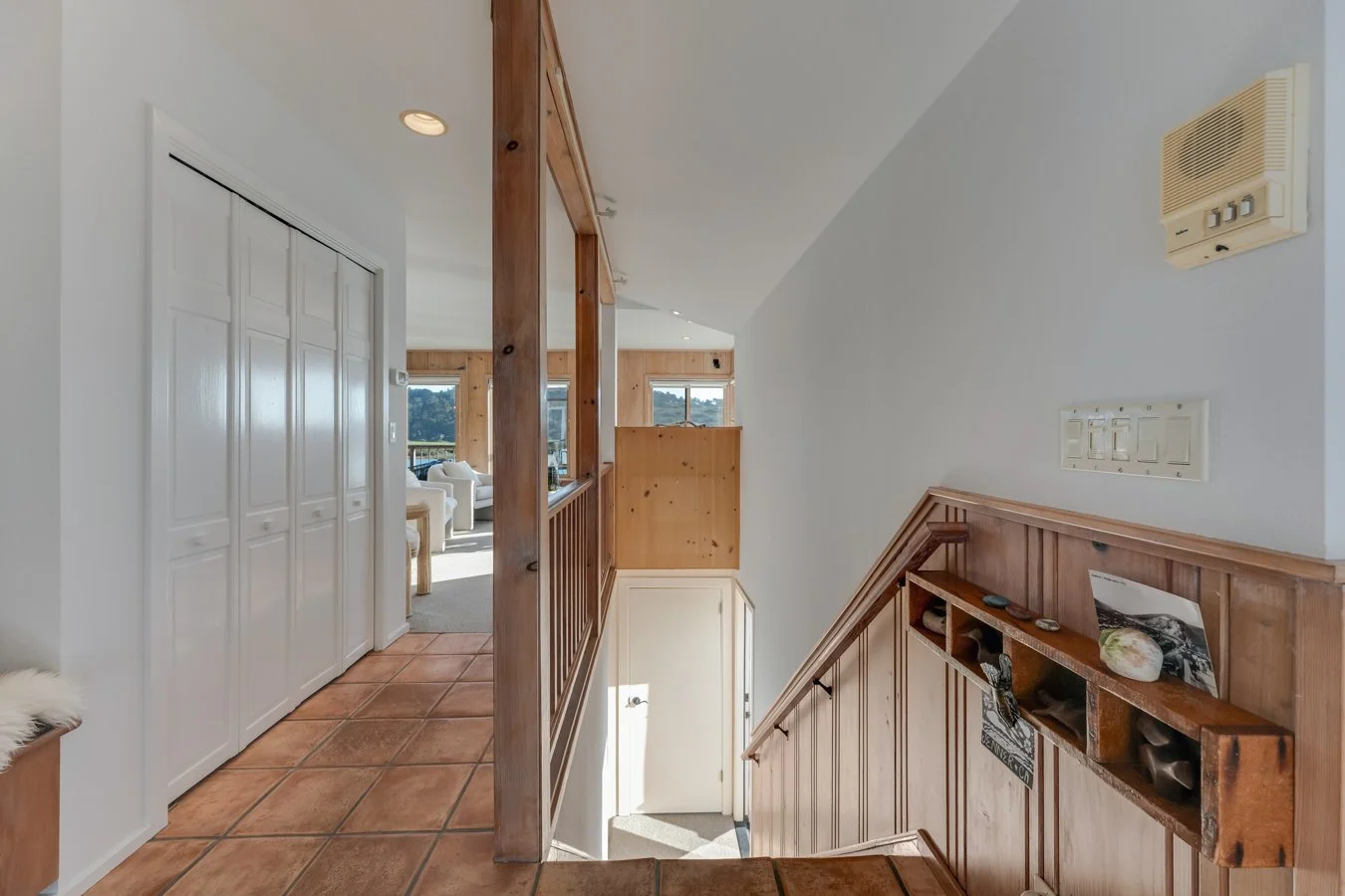 View of a house interior showing a staircase with wooden railing, tiled floor, closet doors, and a living room with windows and furniture in the background.