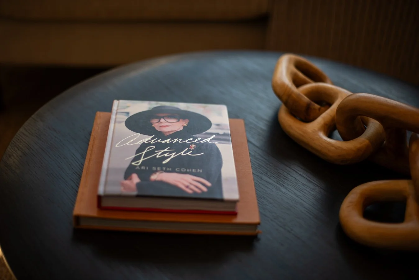 A book titled 'Advanced Style' by Ari Seth Cohen resting on a dark table with a decorative wooden chain link sculpture beside it.