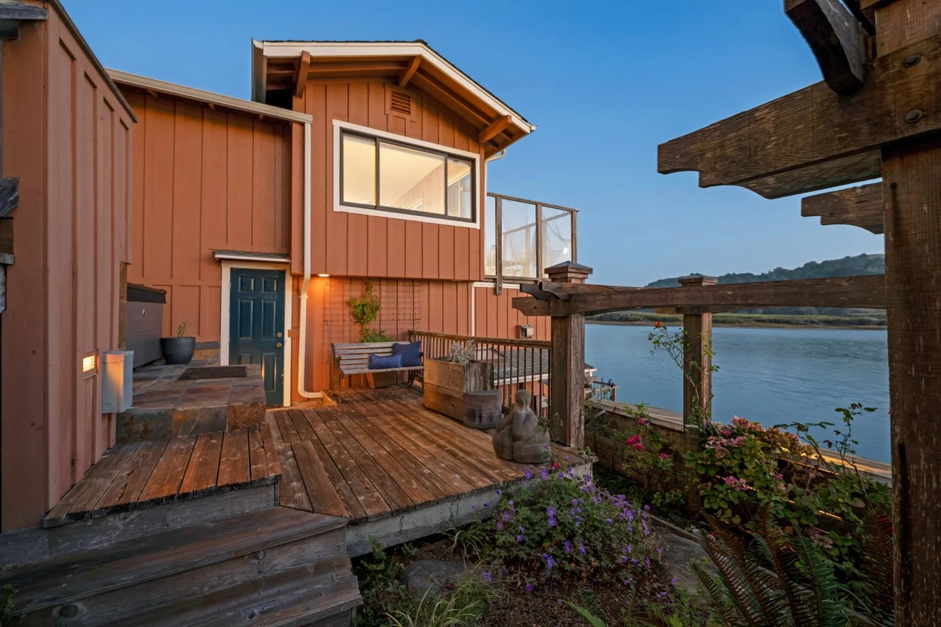 A wooden deck with a bench, potted plants, and a Buddha statue next to a water body with a house in the background during evening.