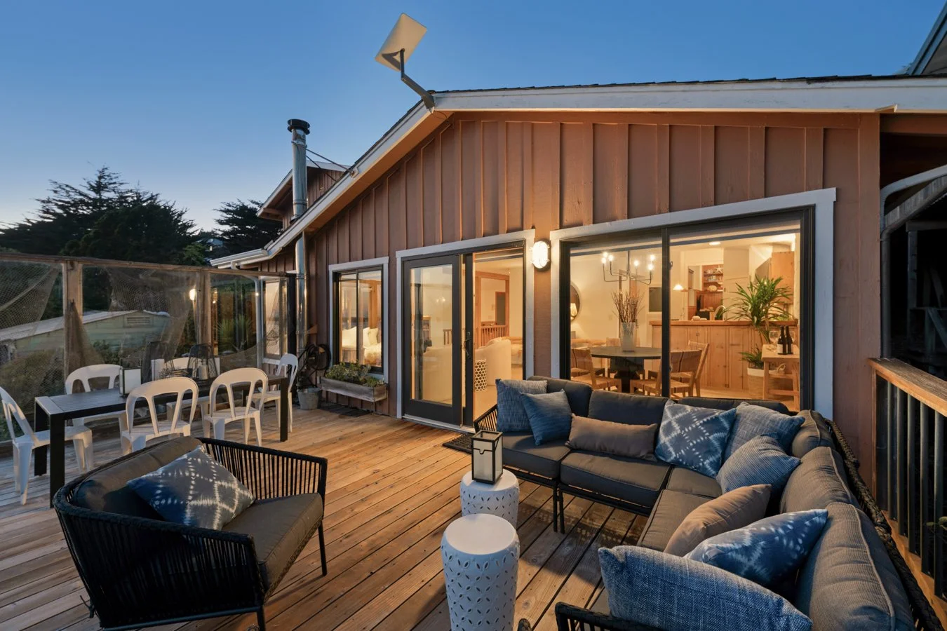 Exterior view of a wooden deck at dusk with outdoor seating, including a black sofa with blue and gray pillows, a black chair with a pillow, a white side table with a lantern, and a dining table with white chairs. The house has large glass sliding do