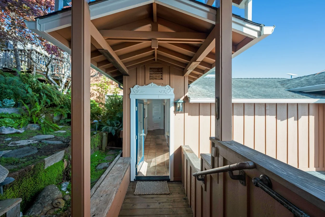 Front porch of a house with a wooden railing, green garden on the left, and a door with a glass window leading inside. Blue sky above.