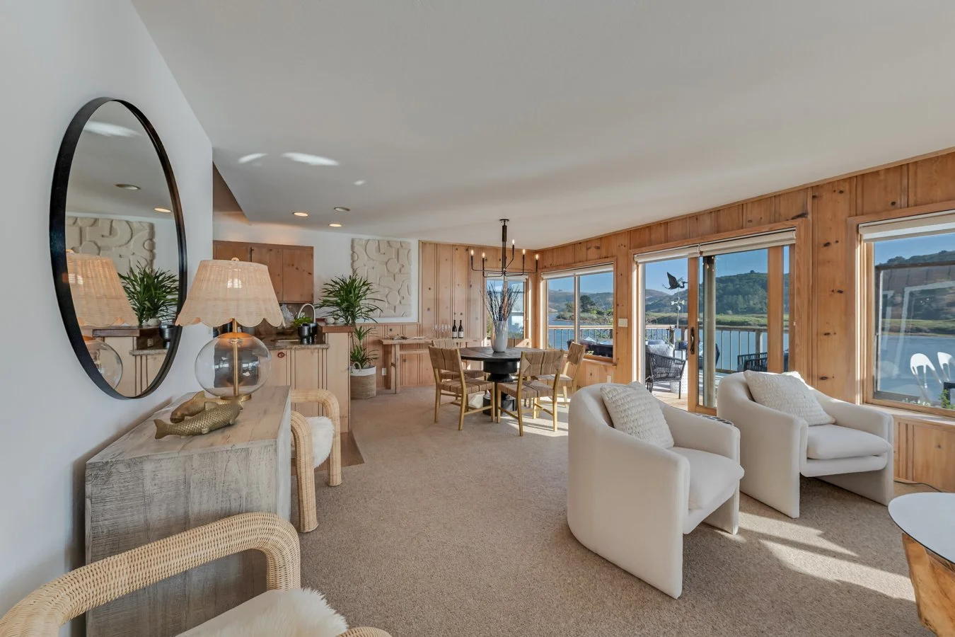 Living room with beige carpet, white armchairs, a round dining table with chairs, large windows with views of water and hills, wood-paneled walls, and sliding glass doors leading outside.