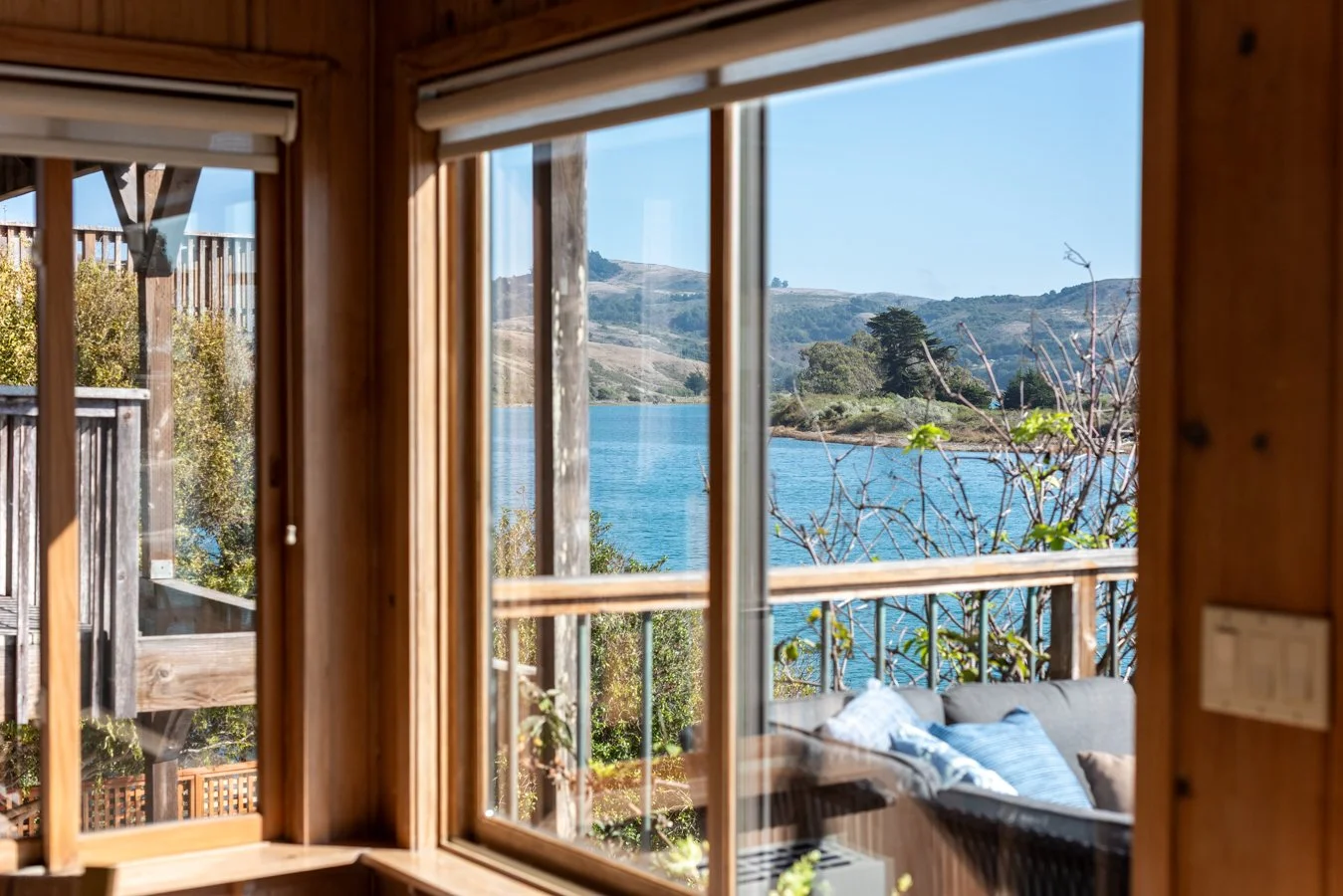 View through a window showing a lake with hills and trees in the background, a balcony with a railing, and part of an outdoor sofa with cushions.