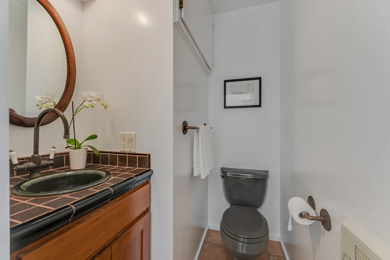 A small bathroom featuring a gray toilet, a wooden vanity with a black sink and bronze fixtures, a mirror, a small potted orchid on the vanity, framed artwork on the wall, a towel ring, and a toilet paper holder.