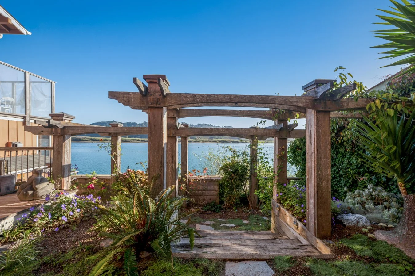 A garden with a wooden arbor overlooking a body of water, surrounded by colorful plants and flowers.