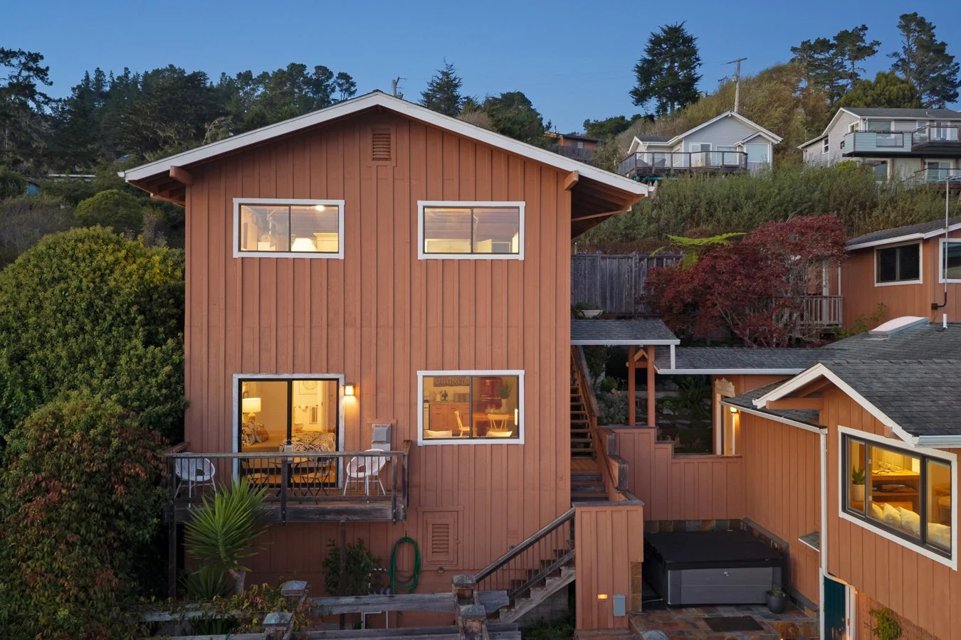 A multi-story wooden house with warm interior lighting, surrounded by green trees and other houses on a hillside, during dusk.