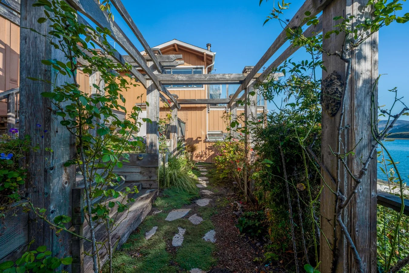 A narrow stone pathway surrounded by green plants and vines leads through a wooden garden trellis towards a house with light brown exterior and large windows, with a body of water visible in the background on the right side under a clear blue sky.