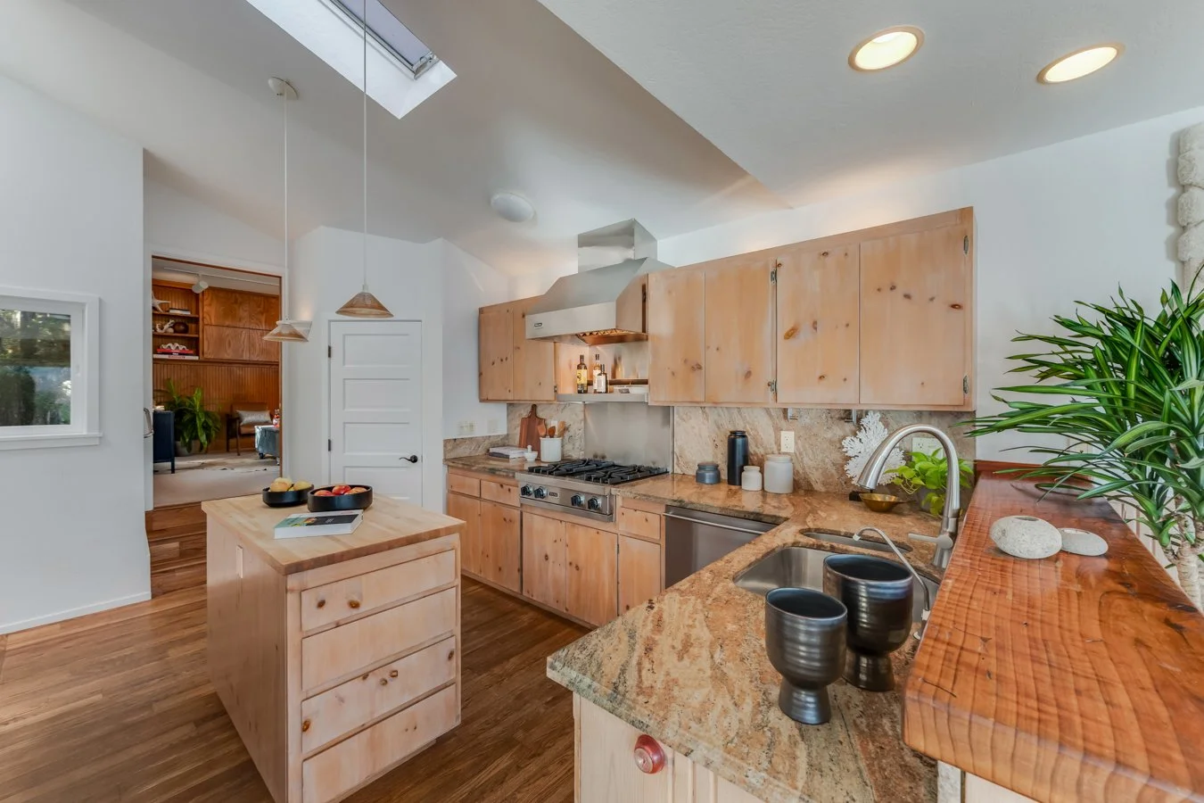 Kitchen with wooden cabinets, granite countertops, a stove, and a small kitchen island. There is a potted plant on the counter and pendant lights hanging from the ceiling. A doorway leads to another room, and a skylight is visible in the ceiling.