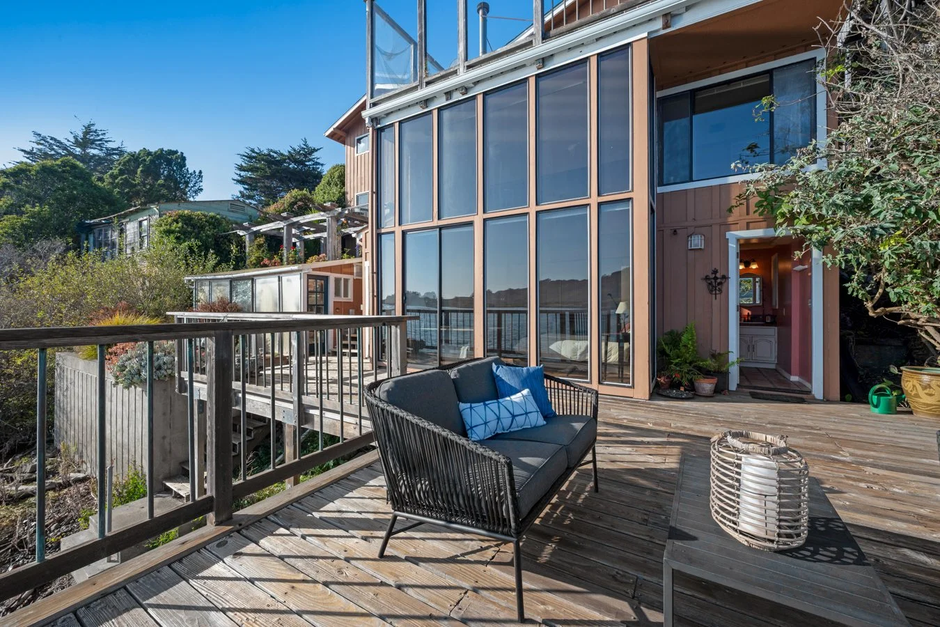 A wooden outdoor deck on a house with large glass windows, overlooking a body of water, with outdoor furniture including a black sofa with pillows and a metal table.