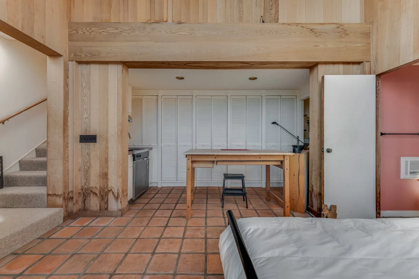 Interior view of a room with terracotta tiled floor, wooden framing, and a kitchenette area with white louvered cabinets, a small sink, and a work table. A black stool and a black desk lamp are present, with partially visible staircase and bed in the