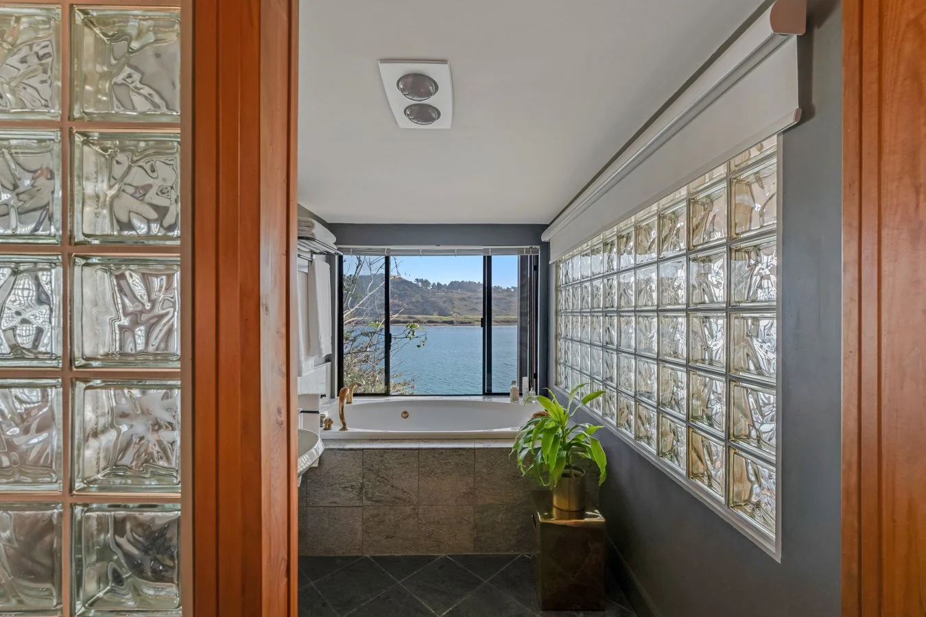 Bathroom with a bathtub overlooking a lake, view through large window, glass block wall, and a potted plant on a stand.