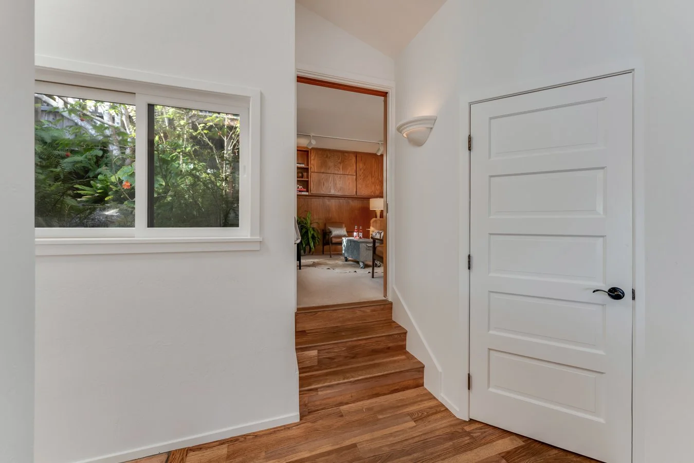 Entryway with a window showing greenery outside, a wooden step, and a door leading to a room with wood-paneled walls, furniture, and houseplants.