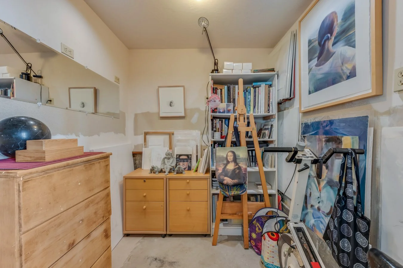 A cluttered interior room with wooden furniture, a bookshelf, framed artwork, and various art supplies and decorations.